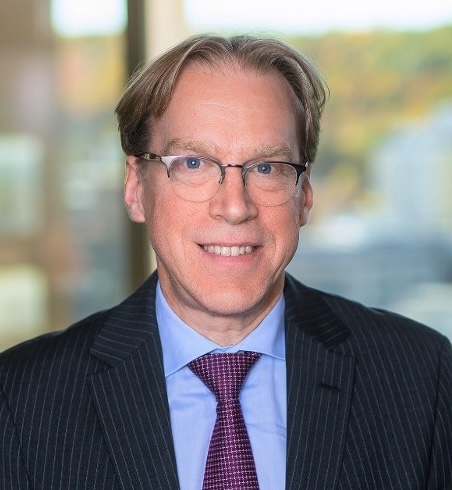 A man with glasses and light brown hair, wearing a dark pinstripe suit, a light blue shirt, and a purple tie, smiles at the camera. The background is blurred, featuring a window with an out-of-focus view outside.