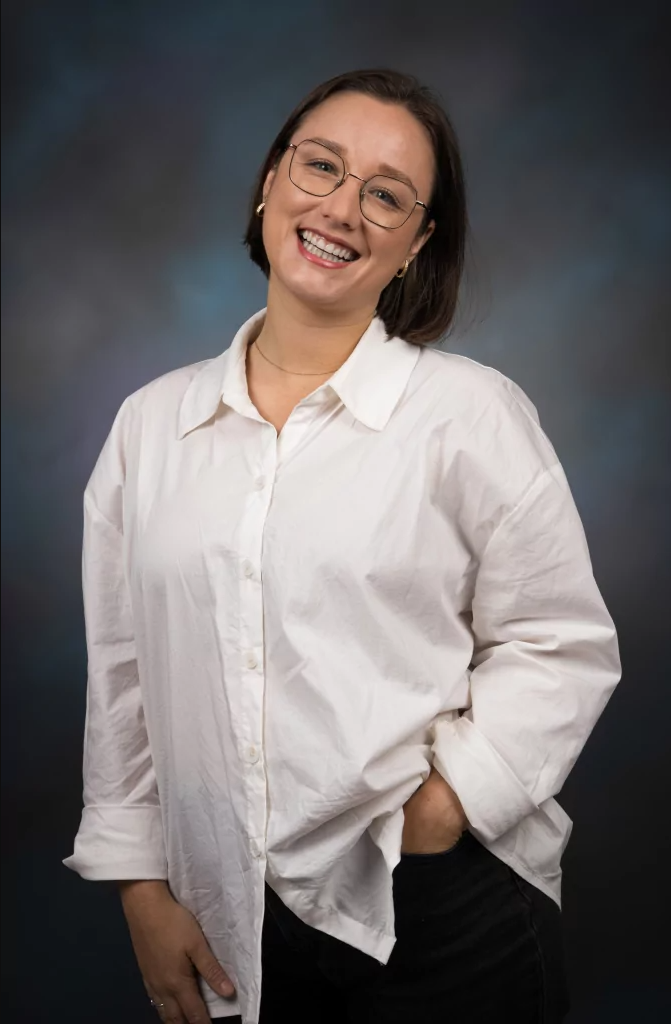 Smiling person with short hair and glasses, wearing a white button-up shirt and dark pants, stands against a dark, cloudy background with one hand in their pocket.