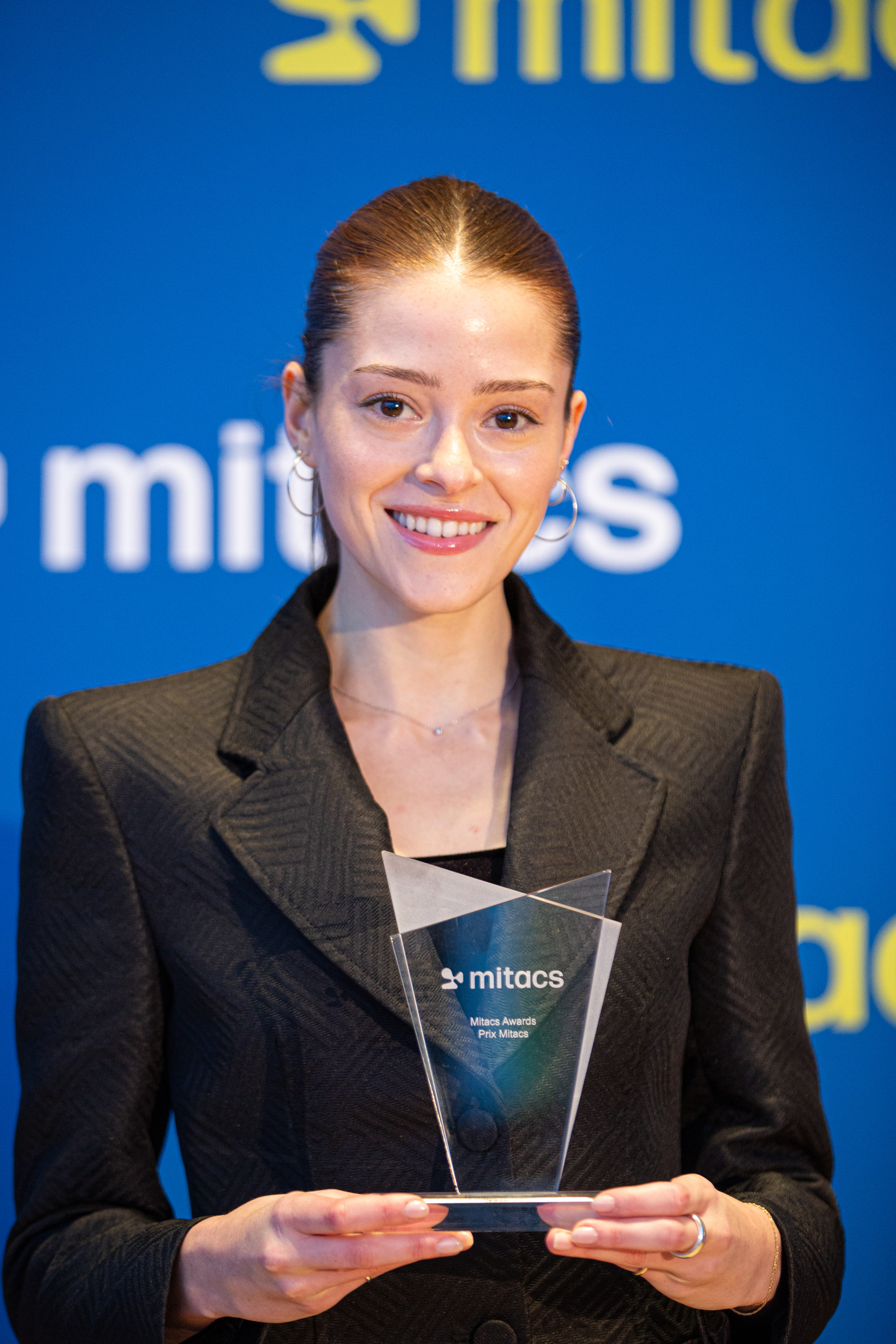 A professional young woman holds a award in front of a Mitacs background.