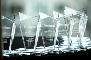 A row of transparent glass awards labeled Mitacs Innovation Awards with names and dates engraved, displayed on a dark table. The background is softly blurred.