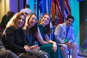 A group of five people sitting and smiling at an event, with various international flags in the background. One person holds a glass. The setting has a lively and social atmosphere.