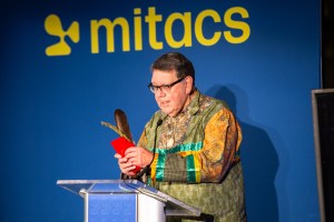 A person stands at a podium against a blue background with the word Mitacs. They are wearing a colorful patterned jacket and holding a feather and a red card, speaking into a microphone.