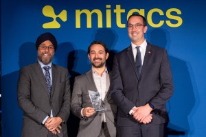 Three men in suits stand in front of a blue backdrop with the Mitacs logo. The man in the middle is holding an award and smiling.