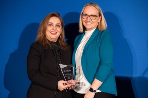 Two women stand smiling against a blue background. The woman on the left, with long hair, holds a glass award. The woman on the right, wearing glasses and a teal blazer, has short blonde hair. Both appear pleased and are facing the camera.