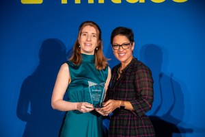 Two women smile while standing in front of a blue backdrop with yellow text. One woman holds a clear award.