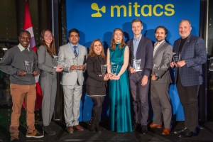 A diverse group of eight people holding awards stand on a stage with a blue Mitacs backdrop and a Canadian flag. They are dressed in formal attire and appear to be at a celebratory event.