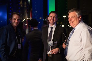 Three men in business attire are smiling and holding awards at a formal event. They stand in a well-lit room with a dark backdrop and other people mingling in the background.